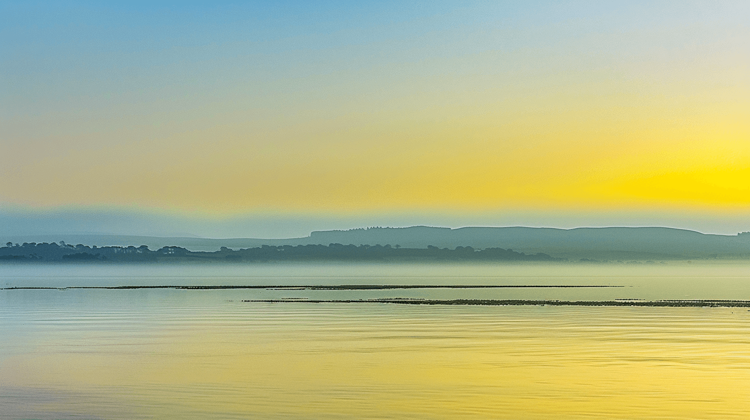 A calm lake at sunrise with soft mist and distant hills